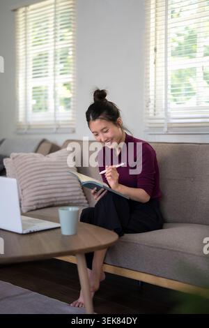 Asiatische Erwachsene weibliche medizinische Fachkraft Schreiben in Journal Notizbuch sitzend auf Couch Laptop Kaffeetasse Fenster Jalousien Stockfoto