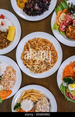Top-down-Komposition mit verschiedenen Tellern und Speisen. Die Teller enthalten Reis, Nudeln, Bohnen, Gemüse, Fleisch und Pommes frites. Warmes, diffuses Licht Stockfoto