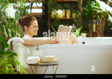 Eine Erwachsene Frau lächelt ruhig in einer weißen Badewanne, vertieft in ein Buch. Umgeben von üppigen grünen Pflanzen und sanftem Tageslicht, ist die Szene capt Stockfoto