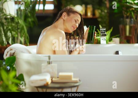 Frau mittleren Alters mit nassbraunem Haar entspannt sich in der weißen Badewanne, Augen geschlossen, Produkt aus dunkelgrüner Flasche auftragen. Grüne Pflanzen und weiche Beleuchtung cre Stockfoto