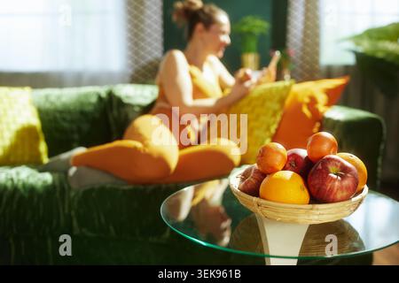 Eine Nahaufnahme eines Korbkörpers mit Äpfeln und Orangen auf einem Tisch, mit einer Frau in orangefarbener Sportswear, die im Hintergrund auf einem grünen Sofa u verschwimmt Stockfoto