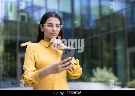 Geschäftsfrau denkt und konzentriert sich, während sie Inhalte auf ihrem Smartphone überprüft und steht mit einem durchdachten Ausdruck außerhalb eines modernen Bürogebäudes Stockfoto
