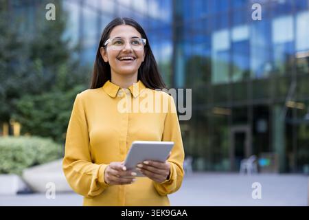Lächelnde Geschäftsfrau, die ein digitales Tablet hält, sieht sich zukünftigen Erfolg und professionelles Wachstum vor, während sie außerhalb eines modernen Bürogebäudes steht Stockfoto