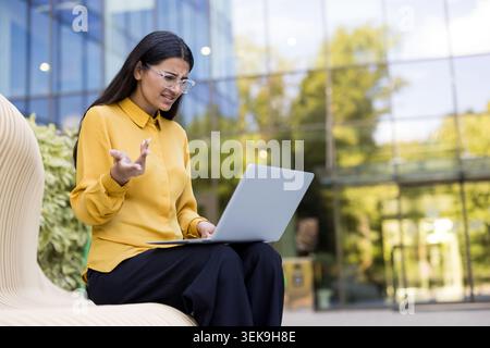 Junge Geschäftsfrau mit Brille, die sich verwirrt und gestresst fühlt, bei der Benutzung ihres Laptops außerhalb eines modernen Bürogebäudes auf ein Problem stößt und frustriert mit Händen Gesten macht Stockfoto