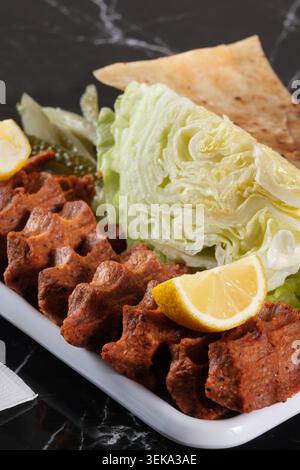 Blick aus einem hohen Winkel auf handgeformte türkische Zigarrenkofte auf einer weißen quadratischen Platte mit geschnittenen Tomaten, Salat, Fladenbrot, Zitrone und Gurken über einem schwarzen märz Stockfoto