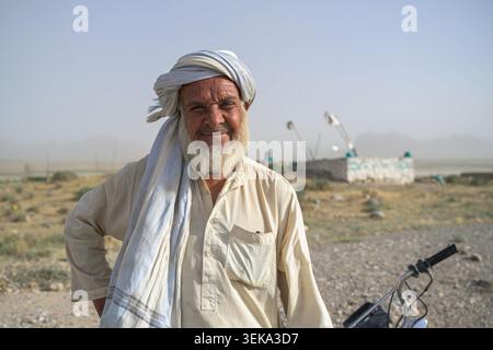 Mann aus kandahar, afghanistan, lächelnd mit weißem Bart und traditioneller Kleidung, selbstbewusst neben einem Motorrad in einem trockenen setti im Freien Stockfoto