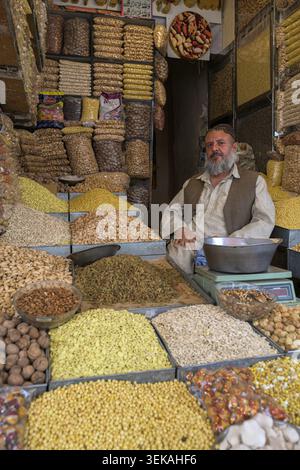 Afghanischer Händler, der zwischen reichlich getrockneten Früchten, Nüssen und verschiedenen Samen in großen Häufchen sitzt und Produkte auf einem geschäftigen traditionellen Markt verkauft Stockfoto