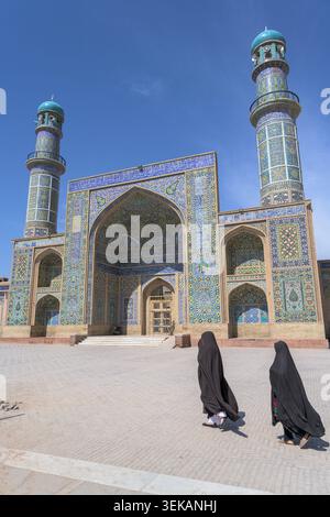 Zwei Frauen in Burka, die vor dem reich verzierten Eingang mit Fliesen und Minaretten der großen Moschee von Herat laufen. Stockfoto