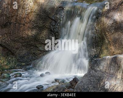 Kleiner Wasserfall, der über strukturierte Felsen in einen flachen Bach in einer friedlichen natürlichen Umgebung stürzt. Stockfoto