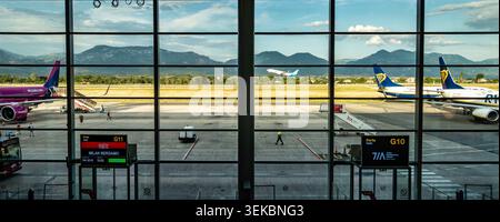 Tirana International Airport in Albanien, durch Terminalfenster gesehen, zeigt Flugzeuge auf dem Asphalt, Gates G10 und G11 und ferne Berge. Stockfoto