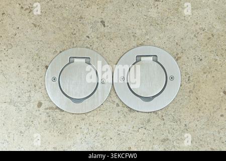 Scharfe und detaillierte Draufsicht auf zwei runde Bodenauslässe aus Edelstahl mit geschlossenen Abdeckungen. Die Metallbuchsen sind bündig in eine strukturierte eingebaut Stockfoto