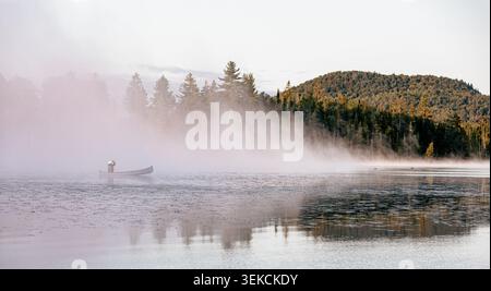 Mann auf einem Boot auf einem See mit dem Morgennebel mit Bäumen auf einem Hügel im Hintergrund Stockfoto