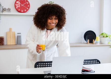 Lächelnde schwarze Frau, die Bademantel trägt, Orangensaft trinkt und in der Küche einen Laptop benutzt Stockfoto