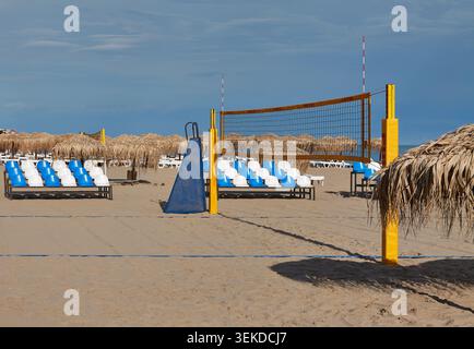 Der Beachvolleyballplatz verfügt über gelbe Pfosten und ein Netz mit Reihen weißer Sonnenliegen und Strohschirmen am Sandstrand unter klarem Himmel Stockfoto