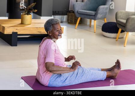 Ältere afrikanische Frau trägt rosa Oberteil, Hosen, lächelt, dehnt sich auf einer lila Yoga-Matte im Wohnzimmer aus Stockfoto