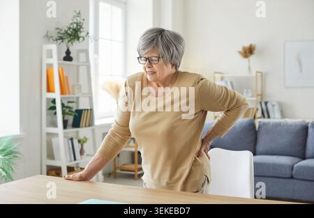 Eine ältere Frau, die zu Hause steht und ihren Rücken berührt, leidet unter Rückenschmerzen. Stockfoto