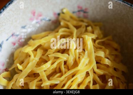 Aus nächster Nähe sehen Sie frische flache gelbe Eiernudeln in einer gemusterten Keramikschale, die für das Kochen einer authentischen asiatischen Mahlzeit bereit ist, die ein appetitliches und hohes Niveau schafft Stockfoto
