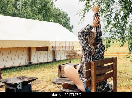 Junge kaukasische Frau mit langen blonden Haaren sitzt auf einer Holzbank und streckt ihre Arme in einem ländlichen Campingplatz mit einem Zelt im Hintergrund. Stockfoto