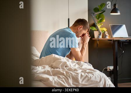 Mann, der auf einem ungebauten Bett sitzt, Hände nahe dem Gesicht im Schlafzimmer. Das Konzept von Stress, Angst, Müdigkeit und seelischem Wohlbefinden zu Hause. Stockfoto