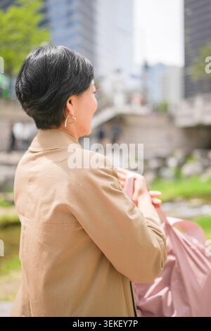 Frau steht und blickt in ruhiger Besinnung auf die Skyline der Stadt. Ihr kurzes, dunkles Haar umrahmt ihr ruhiges Profil. Der weiche, strukturierte Mantel weist auf Komfort und Komfort hin Stockfoto