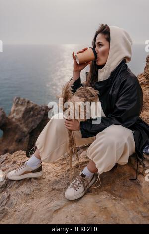Frau Kaffee Ozean. Eine junge Frau im Hoodie trinkt Kaffee, während sie auf einer felsigen Klippe sitzt und das Meer bei Sonnenuntergang überblickt Stockfoto