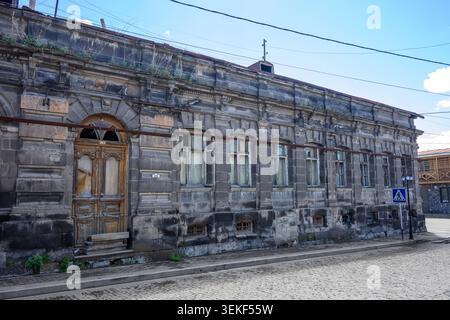 Altes Steingebäude mit bogenförmiger Holztür und mehreren Fenstern. Verwitterte Fassade mit Moos und Rissen sichtbar. Kopfsteinpflaster Straße und Versorgungsleitungen o Stockfoto