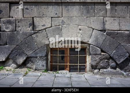Verwitterte Steinmauer mit bogenförmigem Fenster, umrahmt von rauen Blöcken. Holzfenster zeigen Alterserscheinungen. Kopfsteinpflasterboden trifft an abgenutzter Stelle auf Wand Stockfoto