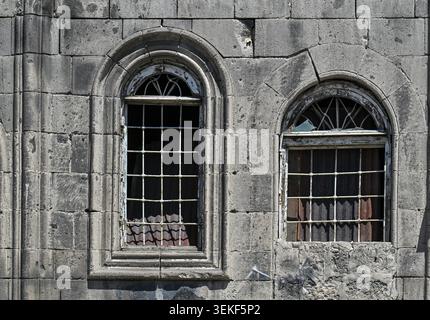 Zwei alte Bogenfenster mit Metallstangen an verwitterter Steinmauer. Fenster zeigen Anzeichen von Verfall und Vernachlässigung. Die Steinstruktur ist rau und gereift. Lichtfilter Stockfoto