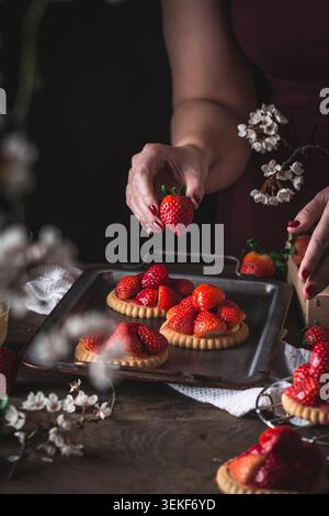 Köstliche, frische, sommerliche Erdbeertörtchen/kleine Kuchen mit einer Frau, die eine Erdbeere im Hintergrund hält Stockfoto