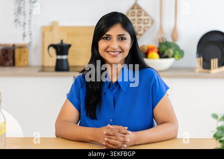 Lächelnde mittelErwachsene indische Frau, die am Tisch in der Küche sitzt Stockfoto