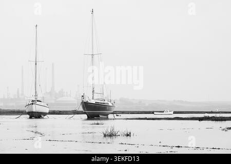 Zwei Segelboote strandeten bei Ebbe in der Angle Bay an der Pembrokeshire Coast. In der Ferne und in Dunst gehüllt, befindet sich die Pembroke Raffinerie. Stockfoto