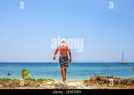 Ein Mann mit langen blonden Haaren läuft unter einem klaren blauen Himmel auf das türkisfarbene Meer zu. Strandurlaub, Sommerschwimmen, tropisches Resort Stockfoto