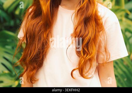 Frau mit langen orangen Haaren steht tagsüber vor großen grünen Blättern in tropischer Umgebung. Mädchen steht draußen. Trägt ein leichtes Hemd Stockfoto