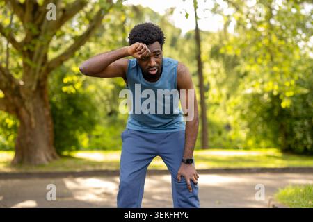 Junger erwachsener Mann, der sich den Schweiß von der Stirn abwischt, sich Atem schnappt und sich während eines intensiven Workouts in einem grünen Park ausruht und körperliche Anstrengung zeigt Stockfoto