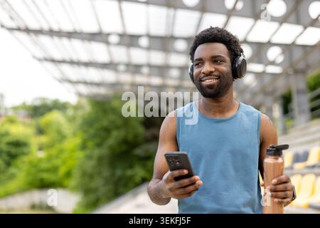 Junger erwachsener Mann, der nach einem Workout mit Wasser versorgt bleibt, Musik mit kabellosen Kopfhörern hört und sein Mobiltelefon im Freien in einem Stadion für einen benutzt Stockfoto