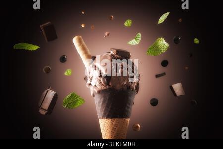 Schokoladeneis in einem in Schokolade getauchten Waffelkegel, umgeben von schwebenden Schokoladenstückchen, Minzblättern und Waffelstab auf dunklem Hintergrund. Stockfoto