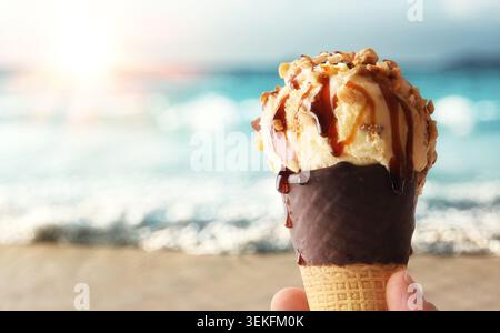 Nahaufnahme von Vanilleeis mit Mandelstückchen und Karamellsauce in einem in Schokolade getauchten Waffelkegel, serviert an einem sonnigen Strand mit verschwommenem Meer Stockfoto