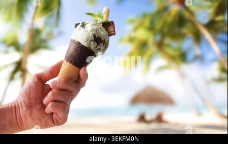 Hand hält Minzschokolade-Eisbecher mit Palmen und tropischem Strand im sonnigen Hintergrund. Stockfoto