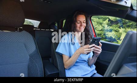 Weibliche Angestellte im Gesundheitswesen lächelt, tippt am Telefon, während sie in einem Fahrzeug sitzt Stockfoto