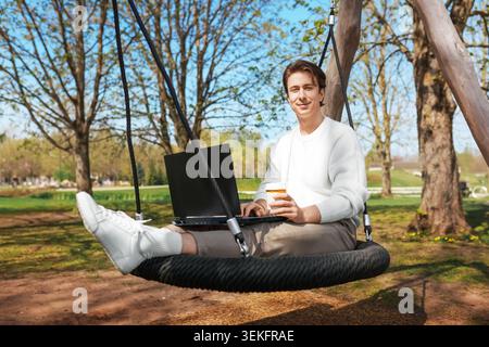 Ein glücklicher Mann genießt Fernarbeit als Freiberufler. Er sitzt draußen auf einer Schaukel im Park mit Laptop und Kaffee. Hochwertige Fotos Stockfoto