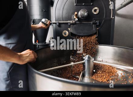 Ein nicht erkennbarer Arbeiter öffnet eine industrielle Kaffeeröstermaschine, um frische Bohnen in ein Fass zu gießen Stockfoto