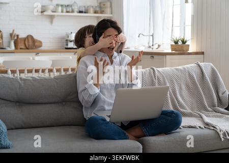 Freiberufliche Frau, die während des Lockdowns auf der Couch im Heimbüro sitzt und am Laptop arbeitet. Spielerisches Kind lenkt von der Arbeit ab, bedeckt die Augen ihrer Mutter, macht Geräusche und bittet die vielbeschäftigte Mutter um Aufmerksamkeit. Stockfoto