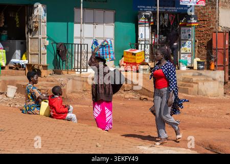 Ugandische Frauen und ein Kind, die sich an Aktivitäten des täglichen Lebens beteiligen, spazieren gehen und sich auf einer lebhaften Straße mit Marktständen und lokalen Unternehmen in der BA ausruhen Stockfoto