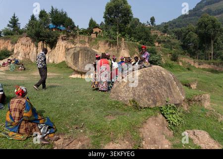 Ugandische Menschen in traditioneller farbenfroher Kleidung engagieren sich in sozialer Interaktion, sitzen und stehen auf und um große Felsbrocken in einem Grün, Stockfoto