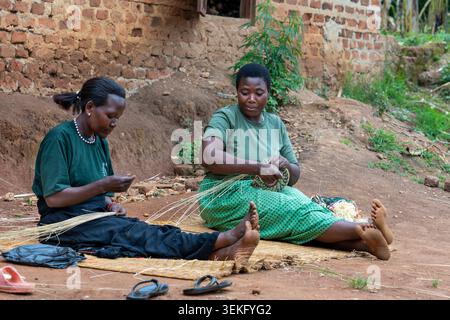 Gruppe afrikanischer Frauen, die auf dem Boden sitzen, lächeln und sich an traditionellem Handwerk des Webenens von Raffiakörben aus natürlichen Materialien beteiligen, umgeben von B Stockfoto