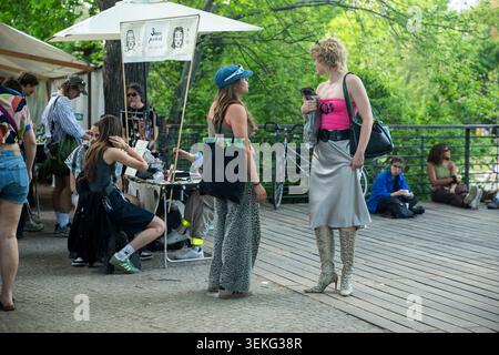 Berlin, Deutschland, 2. Mai 2026. Eine junge Frau mit kurzen lockigen blonden Haaren in rosa Oberteil, silbernem Rock und Schlangenstiefeln spricht mit einer anderen Frau in an Stockfoto