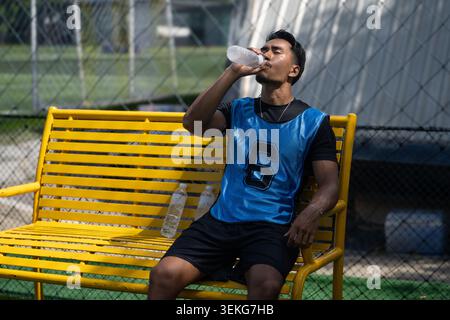 Ein thailändischer asiatischer Fußballspieler trinkt aus einer Wasserflasche, während er sich während einer Pause auf einer gelben Bank neben einem Außenfeld ausruht Stockfoto