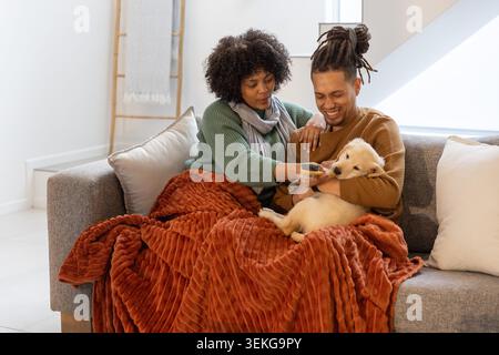 Ein Paar sitzt auf einem grauen Sofa unter dem Oberlicht, trägt Strickpullover, bietet eine Schüssel an und wiegt den Welpen Stockfoto