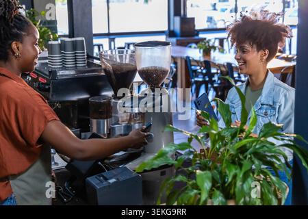 Zwei Frauen, ein Barista, der Espressomaschine benutzt, der Gast, der das Telefon hält, und der POS am Kaffeekocher Stockfoto