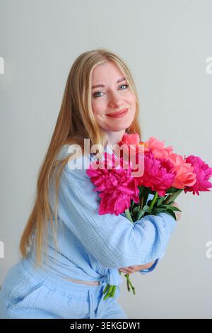 Junge blonde Frau mit frischem Pfingstrosenstrauß in gemütlichem Studio-Setting, modernem Lifestyle und Blumenschönheiten-Fotografie-Konzept. Stockfoto